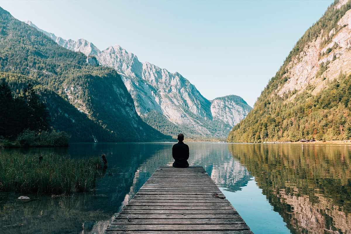 a man meditates at the end of of a pier on a pristine lake surrounded by beautiful mountains